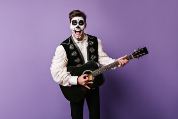 Cheerful man in decorated vest sings song to accompaniment of guitar. Photo of dark-haired guy with skull mask