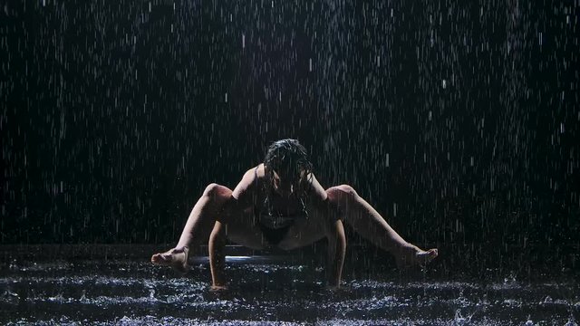 Frog pose performed by a young sportswoman in a dark studio in the rain. Studio light creates shimmer on the surface of the water and the body. Healthy lifestyle concept. Slow motion.