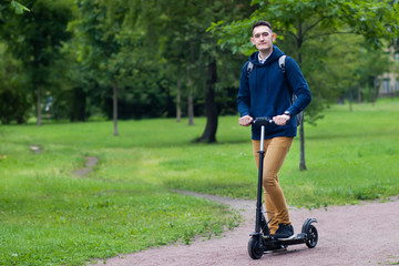 Handsome guy, European man, young college or university student or pupil riding on electric urban modern scooter in summer park with backpack. Eco transport, technology concept. Full length portrait.