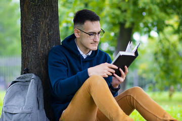 Young concentrated guy in glasses, student reading an interesting book, leaning at a tree, relaxing in summer park with backpack. Handsome smart intelligent man studying outdoors. 