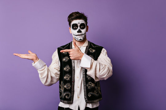 Surprised guy in national Mexican vest pointing finger to left. Portrait of man with painted face with place for tex on lilac background