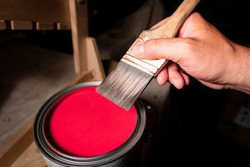 Man's hand prepares to dip paintbrush into one gallon can of red latex paint.