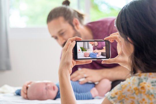 Caucasian woman wife using smartphone taking home video of her family husband with sleeping newborn baby son on the bed. Father and mother spending time together with cute infant child boy at home. 
