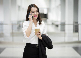 Portrait of a young Asian business woman talking over cellphone and holding cup of coffee in business building. Photo of beautiful girl in casual suite with phone and cup of coffee.