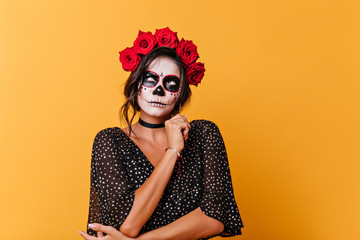 Attractive lady with Halloween makeup looks up thoughtfully. Portrait of girl with red flowers in her hair posing on orange background