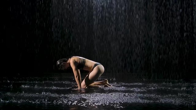 Woman in the rain practices yoga performing asanas. Downward facing dog pose with bent leg. Tightened female body in raindrops. Shot in the studio on a black background. Slow motion.