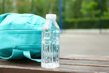 Bottle with water and turquoise backpack on wooden bench outdoors, closeup. Space for text