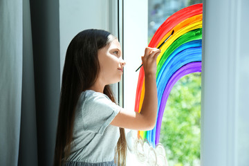 Little girl drawing rainbow on window indoors. Stay at home concept