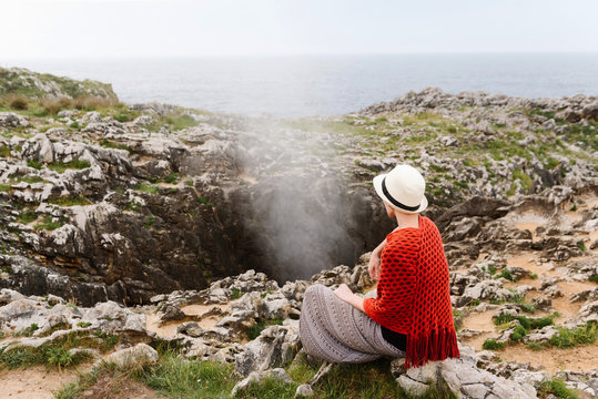 Back view of unrecognizable female traveler in stylish knitwear and hat meditating with mudra gesture while sitting on stone in front of Jesters of Arenillas geyser during trip to Asturian coast of Spain