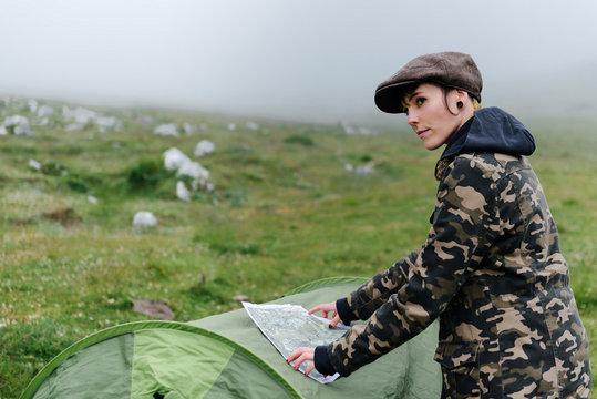 Side view positive female tourist in warm clothes and cap putting map on camping tent and navigating in location while looking away in foggy morning