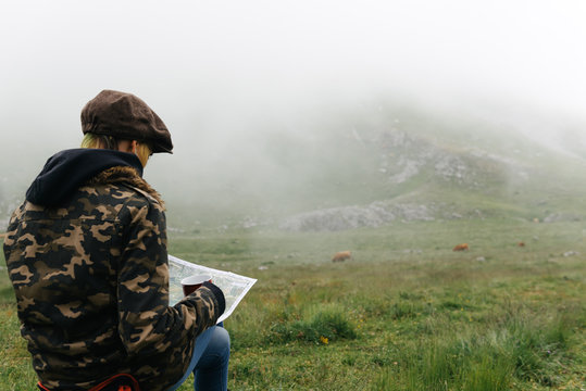 Back view of unrecognizable concentrated female traveler with map sitting with legs crossed on folding chair against camping tent in autumn nature and looking away pensively