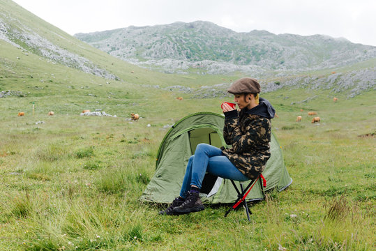 Full length content female traveler in jeans and warm khaki jacket sitting on folding chair near camping tent with cup of hot drink during camping in highlands with closed eyes