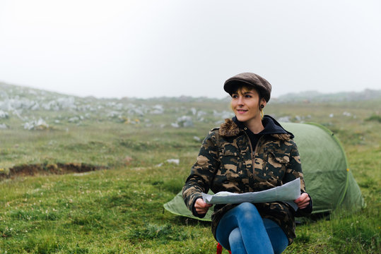 Concentrated female traveler with map sitting with legs crossed on folding chair against camping tent in autumn nature and looking away pensively