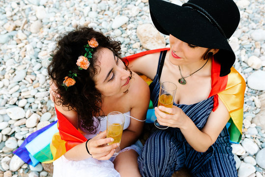 High angle of tender lesbian couple sitting on stony beach and covering with rainbow flag while embracing and clinking glasses with champagne