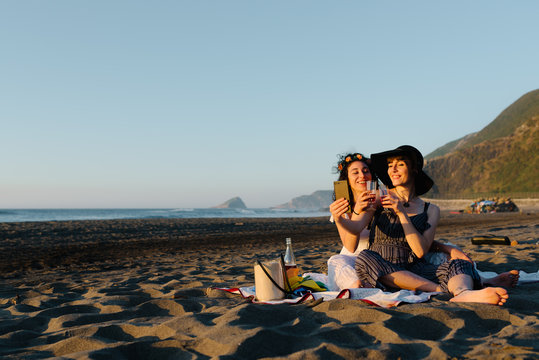 Couple of lesbians resting on seashore with alcohol and taking photo on smartphone while spending vacation together