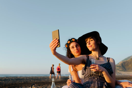 Couple of lesbians resting on seashore with alcohol and taking photo on smartphone while spending vacation together