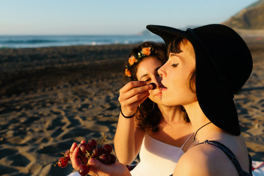 Side view of content woman feeding girlfriend with grape while relaxing on seashore during sunset in summer in back lit