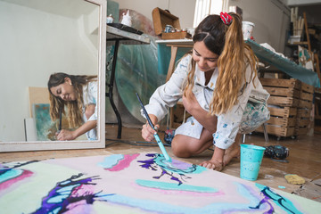 Cheerful barefoot female artist in dirty robe and gloves lying on wooden floor with closed eyes in front of abstract painting in art studio
