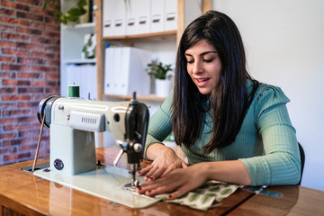 Craftswoman using modern sewing machine while creating soft fabric samples with creative green pattern near lamp in loft style workshop