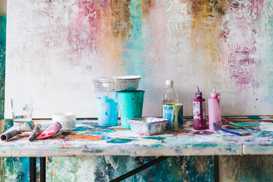 Plastic Bottles With Colorful Paints And Empty Containers Near Artist Tools On Dirty Table Near Multicolored Wall With Uneven Surface In Art Studio