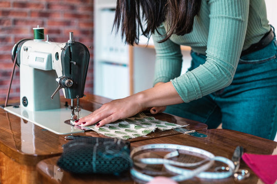 Crop unrecognizable slim female tailor in casual wear with manicure using sewing machine while standing leaned forward near table with soft fabric sample and measuring meter in loft style atelier