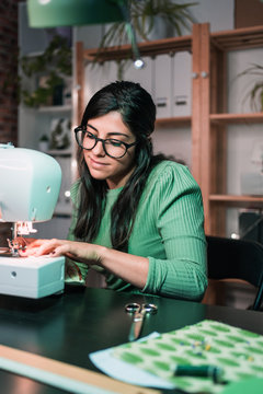 Side view of craftswoman using modern sewing machine while creating soft fabric samples with creative green pattern near lamp in loft style workshop