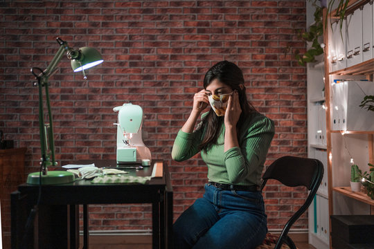 Side view of craftswoman using modern sewing machine while creating masks with creative green pattern near lamp in loft style workshop