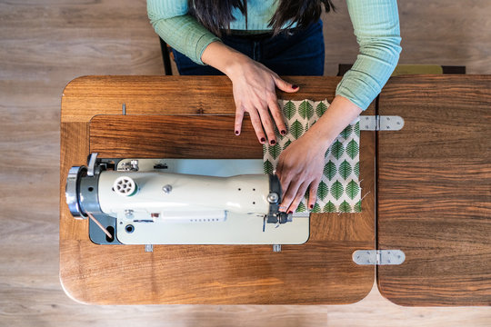 From above unrecognizable craftswoman using modern sewing machine while creating soft fabric samples with creative green pattern near lamp in loft style workshop