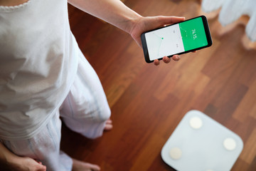From above crop anonymous female in pajama standing on wooden floor near smart digital weight scale and using BMI control application on mobile phone with touchscreen