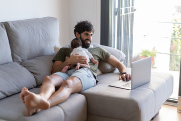 Male freelancer holding baby while lying on sofa and working on project with netbook at home