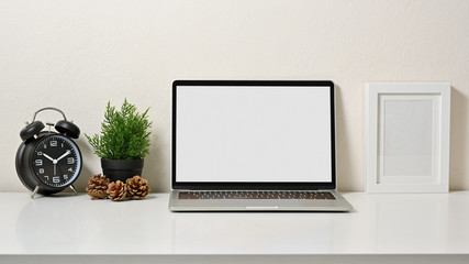 Stylish workspace with laptop, frame pictures, clock, tree and pine cone on white table and white background. Front view.