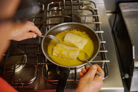From above of crop unrecognizable chef passing fish fillets in melted butter and olive oil in frying pan using fork and spoon while preparing food at home