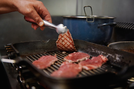 Crop unrecognizable chef frying thin beef slices on grill pan on stove while showing delicious surface of meat using tweezers in kitchen