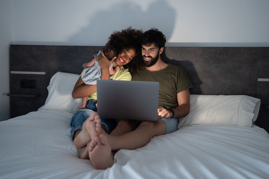 Content Black mother and Arab father sitting on bed with adorable baby and enjoying funny video while using laptop