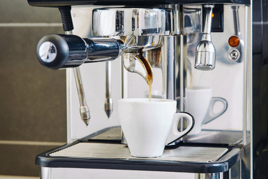 Professional coffee machine made of stainless steel with shiny surface pouring delicious hot coffee into ceramic cup in cafeteria near tile wall