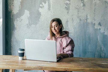 Young female remote employee working on netbook while sitting at wooden table with disposable glass of coffee near old cement wall in flat