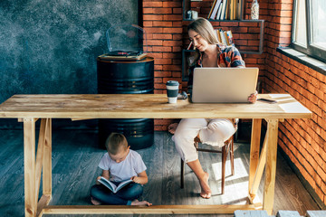 Little child reading book while sitting with crossed legs under wooden table near barefoot woman with netbook and plastic glass of coffee in loft style living room