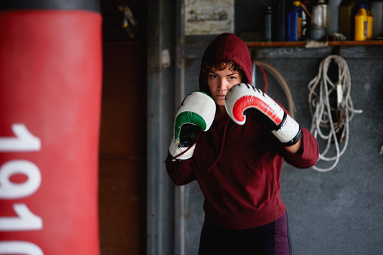 Strong female boxer wearing dark red hoodie and boxing gloves shadows boxing and throwing punches in air while working out in garage gym
