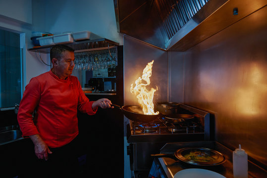 Concentrated chef in uniform holding frying pan while flambeing ingredients in frying pan on slow heat standing in restaurant kitchen in evening and looking at bright fire