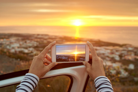Back view of unrecognizable female tourist standing near car and taking photo of amazing sunset over ocean on mobile phone