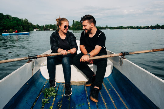 Full body happy smiling couple in black casual outfits rowing boat with oars on calm beautiful pond while looking at each other during clear summer day in Saint George, Transylvania, Romania