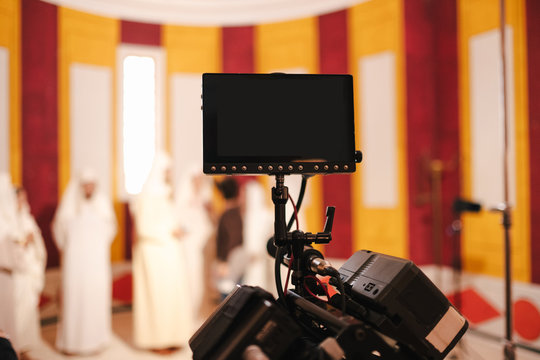 Professional video camera with black display placed against group of people wearing white maxi clothes in creative colorful studio