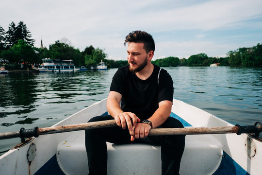 Positive bearded male in casual black outfit rowing boat by using oars on calm picturesque lake and looking away in Saint George, Transylvania, Romania