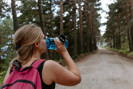 Back view of active young female backpacker standing on pathway among green forest and drinking water while resting during trekking in Navacerrada in Spain