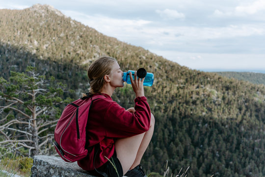 Side view of young female backpacker sitting on stone against green forested hill and drinking water while resting during hiking in Navacerrada in Spain