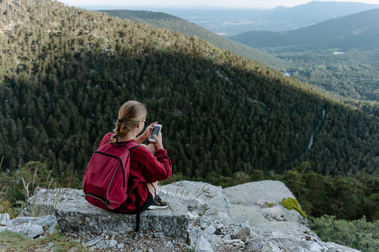 Back view of young female traveler with backpack sitting on stone and checking pictures on smartphone while resting during hiking in picturesque mountains in Navacerrada in Spain