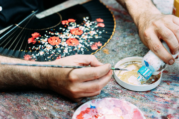 From above crop anonymous male artist pouring white gouache into palette while drawing floral pattern on hand fan in workshop