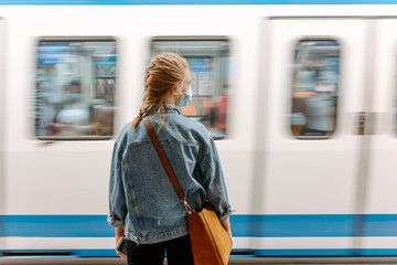 Back view of millennial female passenger in protective mask and casual denim jacket with handbag standing on underground platform in front of moving train