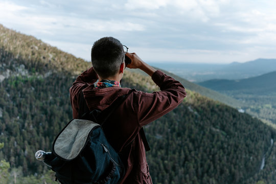 Back view of unrecognizable male tourist with backpack standing on mountain slope and admiring view of green hills while observing landscape with binoculars in Navacerrada in Spain