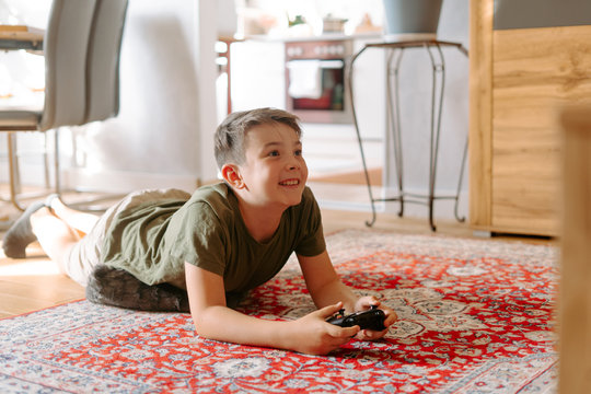 From above of little boy in comfy clothes lying on floor while playing video game with joystick at home